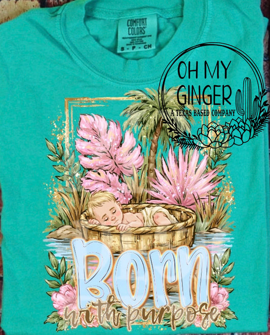 A turquoise short-sleeve t-shirt featuring a graphic print of a baby sleeping in a basket surrounded by pink flowers and palm trees, with the text 'Born with purpose' and a circular 'Oh My Ginger Texas Based Company' logo overlay.