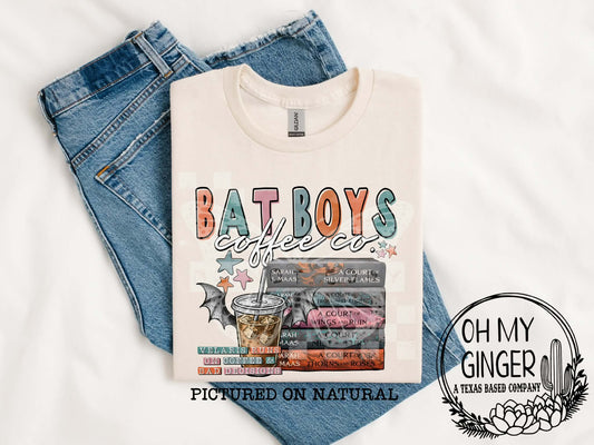 A folded cream-colored graphic T-shirt is laid flat on top of blue jeans against a white fabric background. The shirt features a colorful retro-style 'BAT BOYS coffee co.