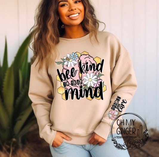 A woman wearing a beige crewneck sweatshirt with a colorful graphic that reads 'bee kind to your mind' surrounded by flowers, paired with light blue jeans, set against a soft-focus background with a potted plant.