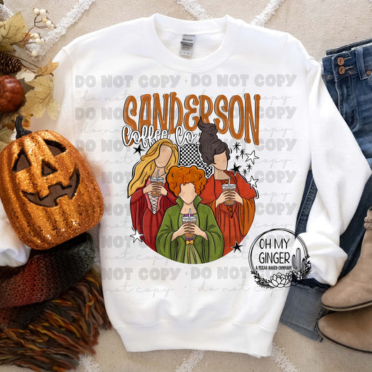 A white crewneck sweatshirt laid flat with a colorful graphic of Sanderson's trio from Hocus Pocus with the text 'SANDERSON Coffee Co.' The sweatshirt is styled with autumn props including a glittery jack-o'-lantern, a knit scarf, jeans, and boots on a patterned beige rug.